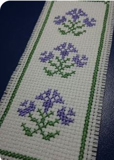 a cross stitch bookmark with purple flowers on white and green trim, sitting on a blue surface