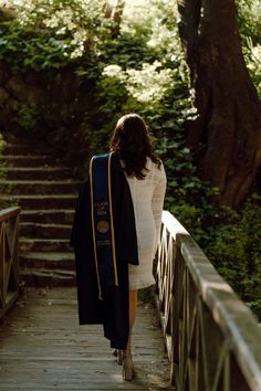 a woman walking across a wooden bridge with a graduation robe over her shoulders and a black stole around her neck