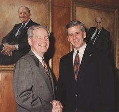 two men shaking hands in front of paintings and smiling at the camera, with one man wearing a suit and tie