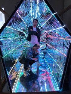 two men are standing in front of an art installation that looks like glass and has colorful lights on it