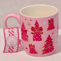 a pink coffee mug with red and gold christmas trees on it next to a small gift bag