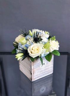 a white and blue flower arrangement in a wooden box on top of a glass table