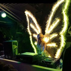 a woman in white dress standing next to a dj booth with lights on her wings