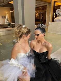 two women dressed in black and white sitting next to each other near a fountain with statues behind them