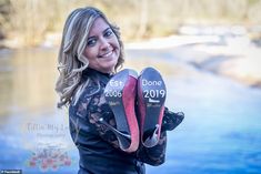 a woman holding two pairs of shoes in front of a body of water with trees behind her