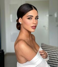 a woman with dark hair wearing a white top and diamond earrings, posing for the camera