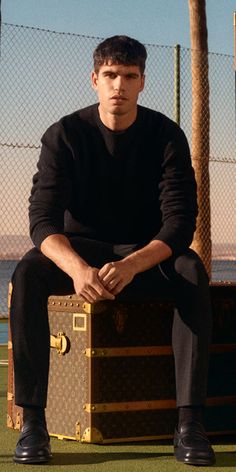 a young man sitting on top of a suitcase next to a tennis court and palm trees