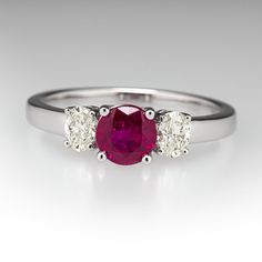 This pretty ring is centered with one (1) round mixed cut natural ruby set into a four-prong setting. The center stone is accented to each side by one (1) oval brilliant cut set into a four-prong setting. The ring measures 5.5mm at the top, rises 5.1mm above the finger, tapering to 2.2mm wide and 1.2mm thick at the base of the shank. This ring is currently a size 6.75. Formal Ruby Three Stone Ring With Diamonds, Luxury Three Stone Ruby Ring, Luxury Oval Three-stone Ruby Ring, Classic Three Stone Ruby Ring With Diamonds, Formal White Gold Ruby Ring With Three Stones, Oval Three Stone Ruby Ring With Diamonds, Oval Ruby Ring With Three Diamonds, Classic Three Stone Round Ruby Ring, Elegant Three Stone Diamond Ruby Ring