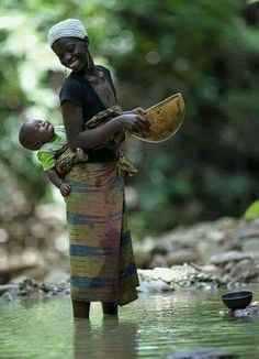 a woman holding a baby in her arms while standing in the water near rocks and trees