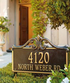 a black and gold sign sitting in front of a house with white flowers around it