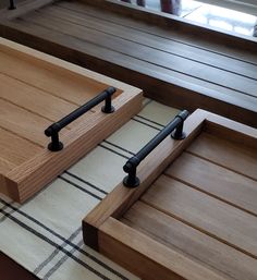 two wooden trays with black metal handles sit on a rug in front of a window