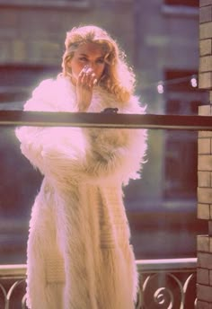 a woman in a fur coat standing on a balcony looking at the camera with her hands to her mouth