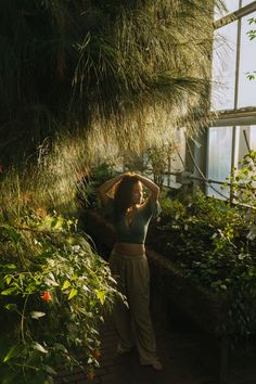 a woman is standing in front of some plants and looking at the camera with her hand on her head