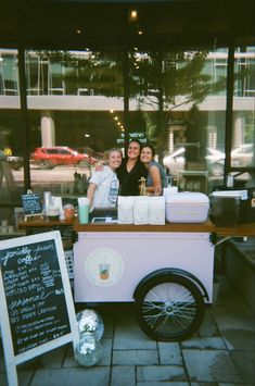 two women standing behind a food cart with drinks on the side and chalkboard menus