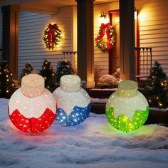 three christmas lights sitting on top of snow covered ground in front of a house with wreaths