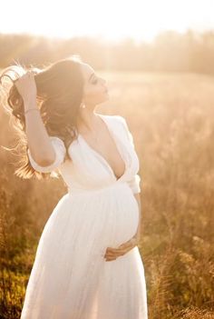 a pregnant woman in a white dress standing in a field with her hands on her head