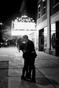 a man and woman kissing on the sidewalk in front of a movie theater at night