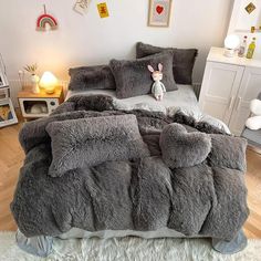 a bed covered in grey fluffy blankets and pillows with a dog sitting on top of it