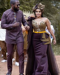 a man and woman dressed in african attire walking down a dirt road with people behind them