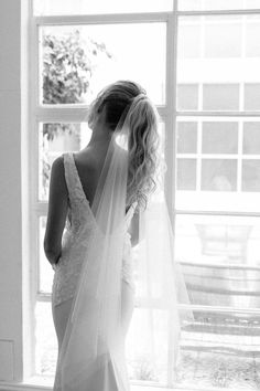 a woman standing in front of a window wearing a wedding dress with a long veil