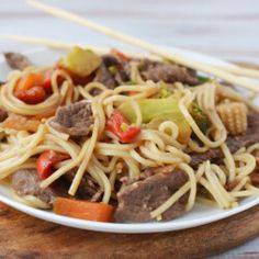 a white plate topped with noodles and meat next to chopsticks on a wooden board