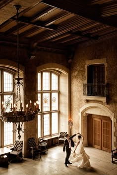 a bride and groom dancing in an old - fashioned room with chandeliers, windows, and fireplace