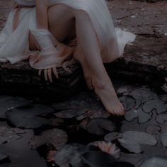 a woman in white dress sitting on rock next to water with her legs crossed and head down