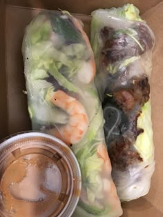 there are two rolls with shrimp and lettuce wrapped in plastic wrappers next to a cup of dipping sauce