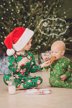 two babies sitting on the floor in pajamas and santa hats eating cookies next to a christmas tree