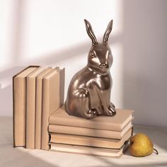 a metal rabbit sitting on top of books next to an apple and pear, in front of a white wall