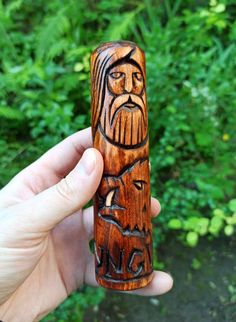 a hand holding a carved wooden object in front of some trees and bushes with green foliage