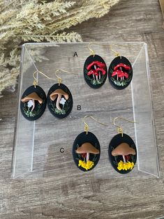 four earrings with mushrooms on them sitting in front of a wooden table next to a plant