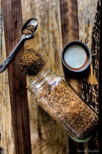 a jar filled with spices next to a spoon on top of a wooden table covered in dirt