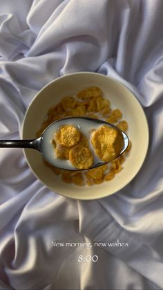 a white bowl filled with cereal on top of a bed next to a silver spoon