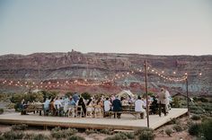 a group of people sitting around a wooden table in the middle of mountains with string lights strung over them