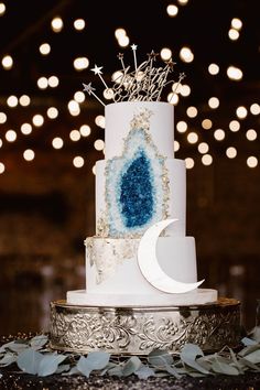 a white and blue wedding cake sitting on top of a silver platter with lights in the background