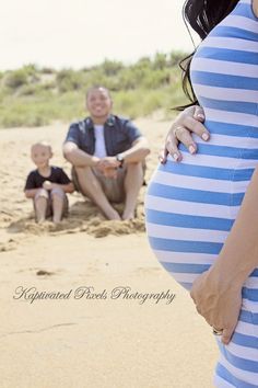 a pregnant woman standing in the sand with her husband and two sons sitting behind her