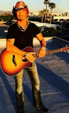 a man holding an acoustic guitar while standing on top of a parking lot with palm trees in the background