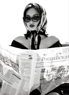 a woman sitting down reading a newspaper with her head wrapped around her neck and wearing sunglasses