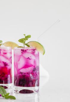 two glasses filled with purple and green drinks next to lemon wedges on a white surface