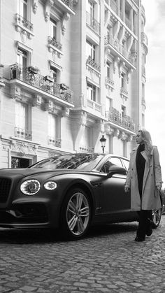 a woman standing next to a black car on a cobblestone street in front of tall buildings