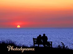 two people sitting on a bench watching the sun set over the ocean in front of them