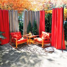 an outdoor patio with red curtains and two orange chairs in front of a small table