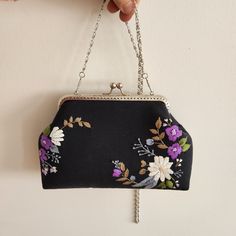 a hand holding a black purse with purple and white flowers hanging from it's chain