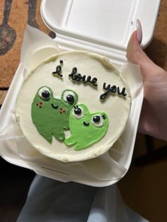 someone is holding up a cake decorated with two green frogs and the words i love you