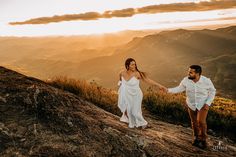 a man and woman holding hands while standing on top of a mountain with the sun setting behind them