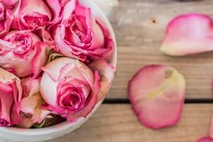 a white bowl filled with pink roses on top of a wooden table next to petals