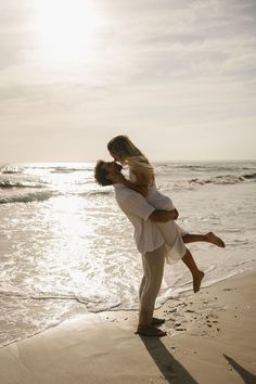 a man holding a woman while standing on top of a beach next to the ocean