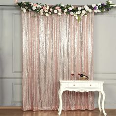 a white table sitting in front of a pink curtain with flowers on it and a vase next to it