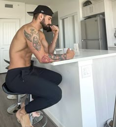 a shirtless man sitting at a kitchen counter talking on a cell phone and drinking from a cup